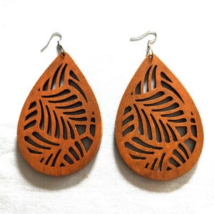 May include: A pair of teardrop-shaped wooden earrings featuring a laser-cut leaf pattern. The earrings are made of a medium-brown wood and are attached to silver-toned metal fishhook ear wires for hanging. The design is symmetrical and open.