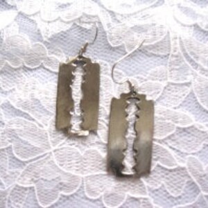 May include: A pair of silver earrings shaped like razor blades. The earrings are hanging from a white lace background.