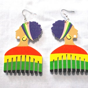 May include: A pair of wooden earrings shaped like a woman's head with a comb design. The earrings are painted in red, yellow, green, purple, and black.