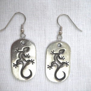 May include: Silver dangle earrings with a lizard design. The lizard is in a dark silver color and is embossed on a silver rectangle.