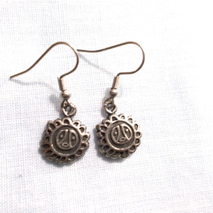 May include: A pair of silver-toned earrings. Each earring features a sun-like design with a central emblem. The earrings have a hook closure and are displayed against a white background. The earrings are likely made of metal.