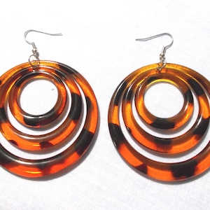 May include: A pair of large, retro-style earrings. Each earring features three concentric circles in a tortoiseshell pattern of amber and black. The earrings have silver-colored fish hook ear wires.