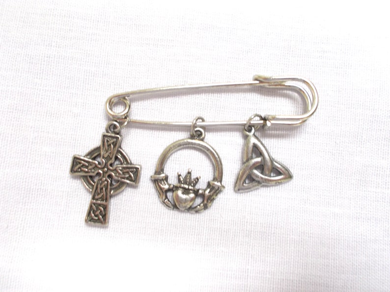 May include: Silver safety pin with three charms: a Celtic cross, a Claddagh ring, and a Celtic knot.