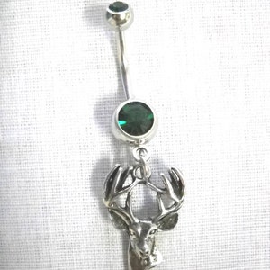 May include: A silver-colored belly button ring with two green gemstones and a deer head pendant. The pendant is a detailed depiction of a deer's head with antlers. The gemstones are round and faceted.