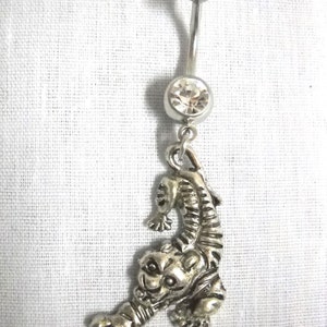 May include: Silver-toned belly button ring with a dangling tiger charm. The ring features a clear, round gem and a curved barbell design. The tiger charm is detailed with stripes and a textured finish, adding a unique touch to this body jewelry.