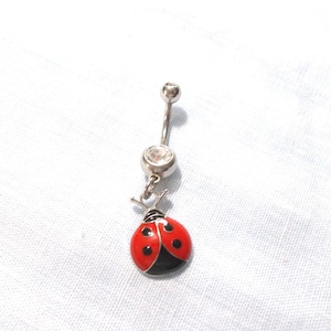 May include: A silver-toned belly button ring featuring a curved barbell with a clear round gemstone at the base. A dangling charm shaped like a ladybug with a bright red body, black spots, and a black head hangs from the bottom of the gemstone.