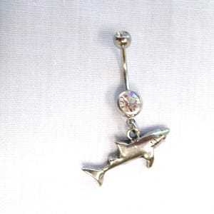 May include: A silver-toned belly button ring featuring a curved barbell with a small ball at the top and a larger, clear, round faceted crystal stone above a dangling, three-dimensional shark charm. The metal has a polished, reflective finish.
