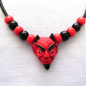 May include: A devil-themed necklace featuring a red devil pendant with black horns and a goatee. The necklace has alternating red and black beads strung on a black cord. The devil's face has a sinister expression.