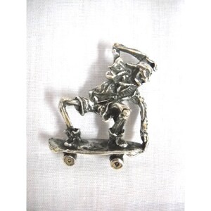 May include: A silver skeleton figurine on a skateboard. The skeleton is wearing shorts and has its arms raised above its head. The skateboard has wheels.
