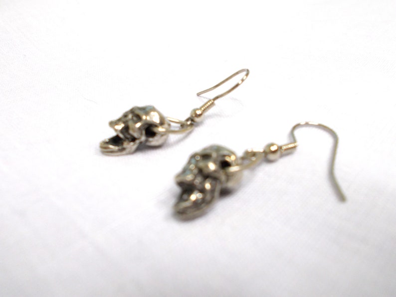 May include: A pair of silver skull dangle earrings. The skulls are detailed and have an open mouth.