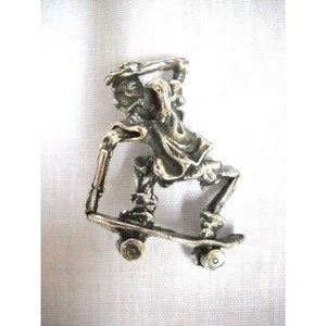 May include: A silver-colored pendant depicting a skeleton skateboarding. The skeleton is wearing a shirt and pants, and the skateboard has four wheels.