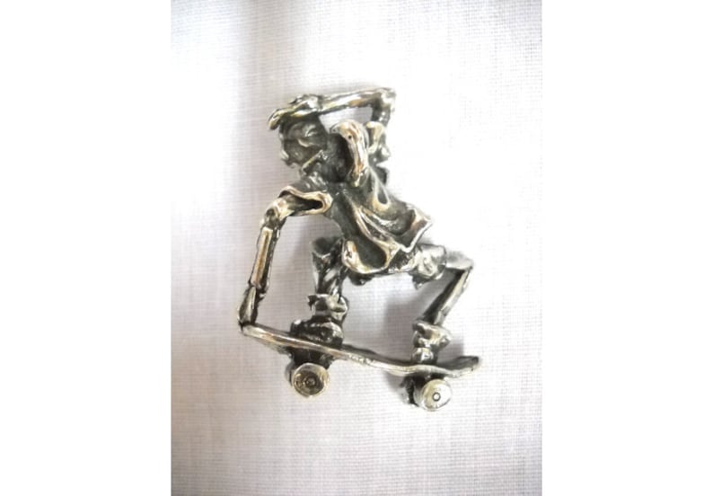 May include: A silver-colored pendant depicting a skeleton skateboarding. The skeleton is wearing a shirt and pants, and the skateboard has four wheels.