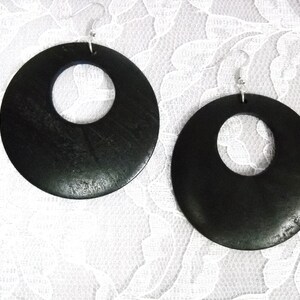 May include: A pair of large, black, round wooden earrings with a circular cutout in the center. The earrings are hanging from silver hooks.