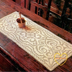 Puede incluir: Camino de mesa crema de tejido abierto con un intrincado patrón floral y de remolinos repetitivo. Un pequeño mortero y maja de latón se encuentran sobre el camino de mesa. El camino de mesa se exhibe sobre una mesa de madera oscura.