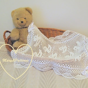 May include: A handmade white crochet baby blanket with a floral pattern, draped over a wicker basket. A tan teddy bear sits in the basket. The blanket has a scalloped edge and white ribbon bows. The text "Mary & Patterns" is in the lower left corner.