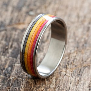 May include: A titanium ring with a colourful wood inlay. The inlay features stripes of black, yellow, orange, and red wood.