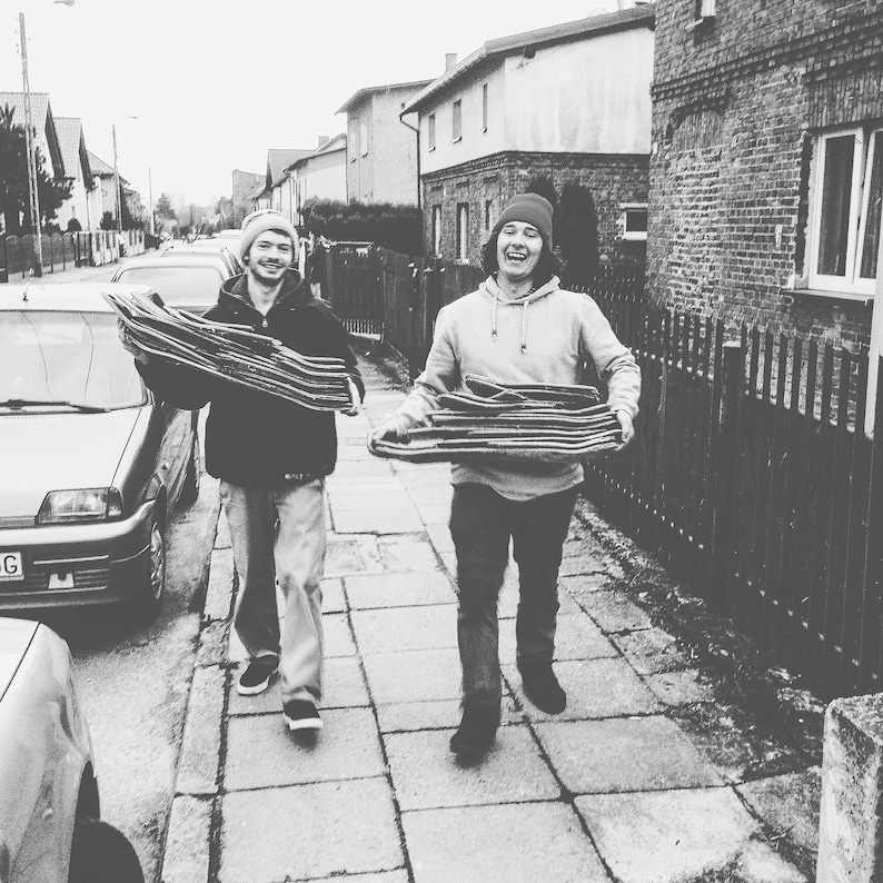 May include: Two people walking down a sidewalk carrying stacks of skateboards. The person on the left is wearing a black jacket and jeans. The person on the right is wearing a grey hoodie and jeans. Both people are wearing hats.
