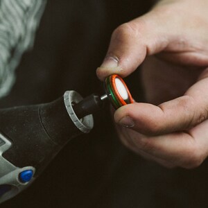 May include: A close-up of a person holding a small, round, multi-colored wooden disc. The disc is attached to a small metal rod, which is connected to a black and gray power tool.