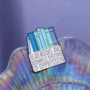 May include: A white enamel pin with a stack of blue and green books and the text "Read Books and Dismantle Systems of Oppression".