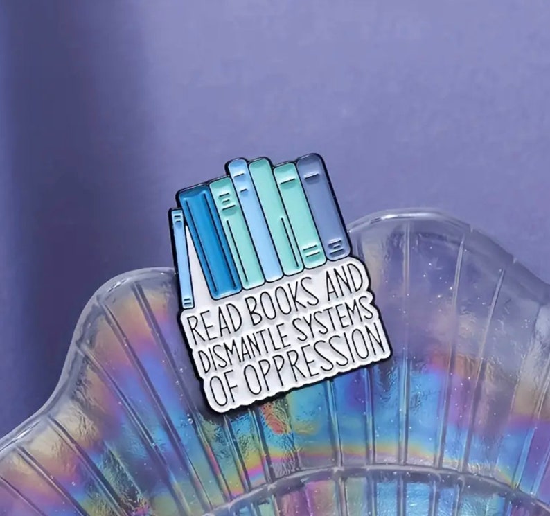 May include: A white enamel pin with a stack of blue and green books and the text "Read Books and Dismantle Systems of Oppression".