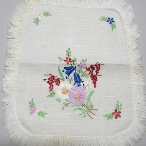 Puede incluir: Un posavasos cuadrado de lino blanquecino con borde de flecos. El posavasos presenta un diseño floral bordado en tonos rojos, azules, morados y blancos. Hojas y tallos verdes acentúan las flores.