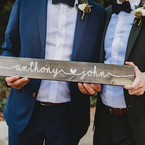 May include: A rustic wooden sign with the names "Anthony" and "John" in white script, separated by a heart symbol. The sign is held by two people, likely at a wedding or engagement event. The sign is a dark gray color.