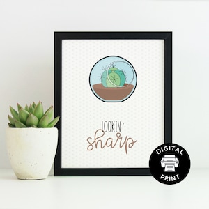 May include: A printable digital art print with a black frame. The print features a cartoon illustration of a succulent in a glass sphere with the text "Lookin' sharp" in brown lettering.