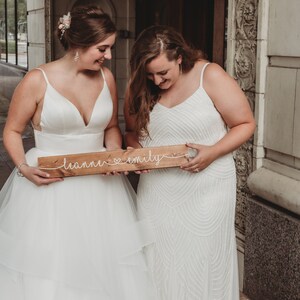 May include: Two people wearing white wedding dresses hold a wooden sign with the text "leanne &hearts; emily" written in white string.