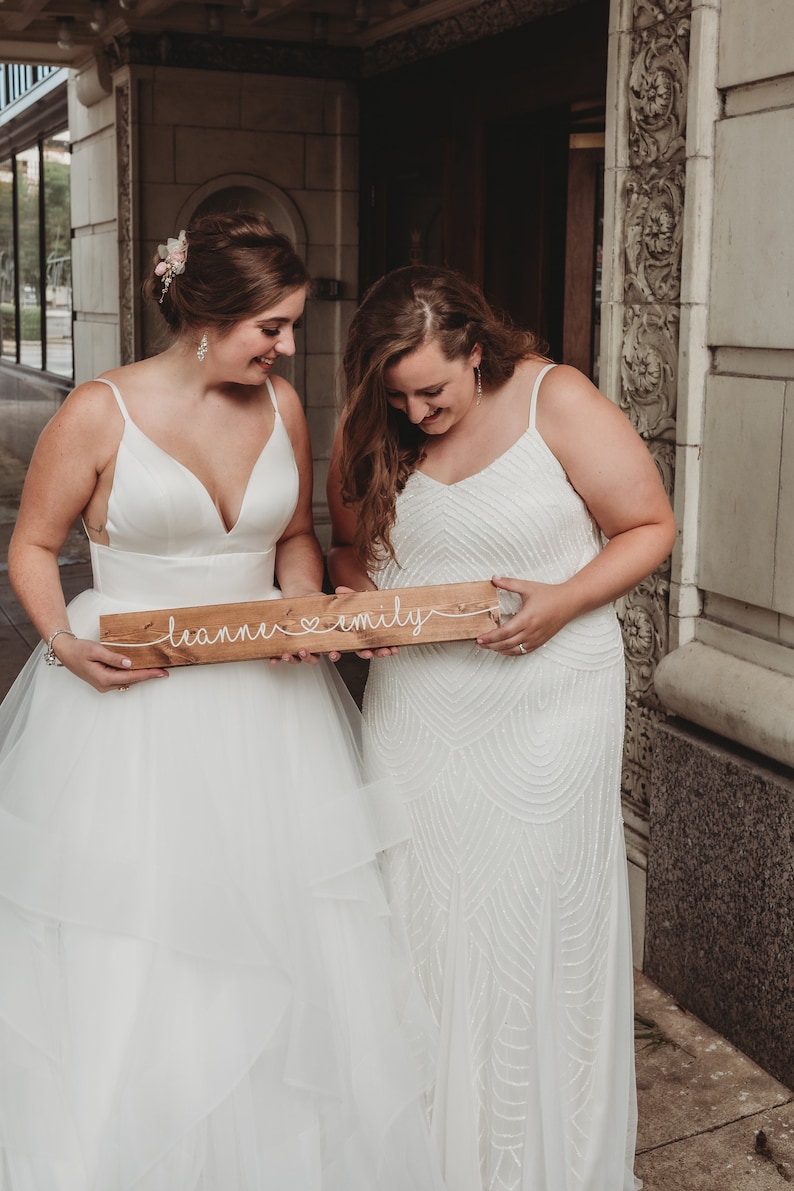 May include: Two people wearing white wedding dresses hold a wooden sign with the text "leanne &hearts; emily" written in white string.