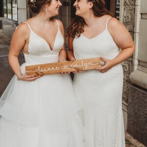 May include: Two people in white wedding dresses holding a wooden sign that says "leanne &hearts; emily".