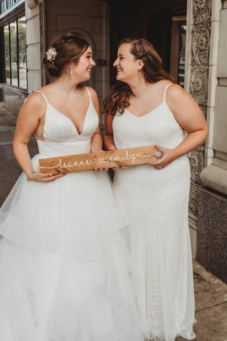 May include: Two people in white wedding dresses holding a wooden sign that says "leanne &hearts; emily".