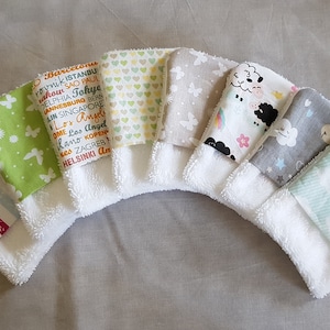 Puede incluir: Conjunto de siete toallitas blancas con cuadrados de tela coloridos cosidos. Los cuadrados de tela presentan varios patrones, incluyendo mariposas, corazones, nubes y un horizonte de ciudad.