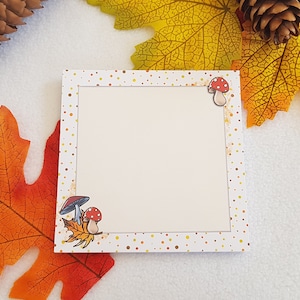 Può includere: Un biglietto bianco con un bordo di pois marroni e arancioni. Il biglietto ha un centro bianco con un disegno di due funghi e una foglia. Il biglietto è circondato da foglie autunnali.