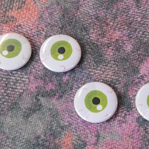 Könnte beinhalten: Vier runde weiße Knöpfe mit grünen Augenmotiven. Die Knöpfe haben einen weißen Hintergrund mit roten Adern und einer schwarzen Pupille. Die Knöpfe befinden sich auf einem pink- und grauen gemusterten Stoff.