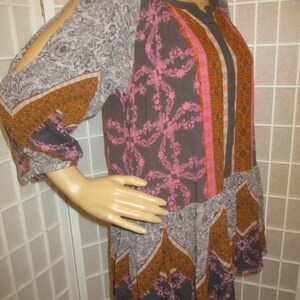 May include: A patterned tunic dress with long sleeves and cut-out shoulders. The dress features a mix of floral and geometric patterns in gray, pink, and orange hues. The dress is displayed on a mannequin.