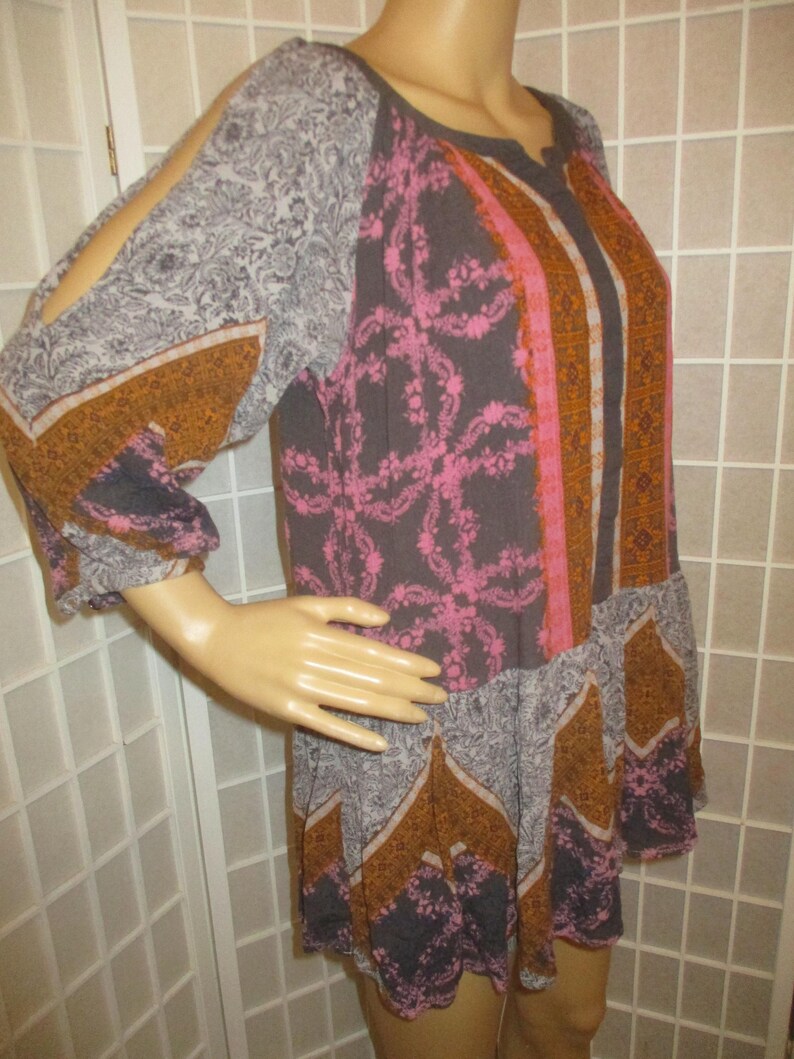 May include: A patterned tunic dress with long sleeves and cut-out shoulders. The dress features a mix of floral and geometric patterns in gray, pink, and orange hues. The dress is displayed on a mannequin.