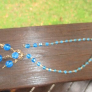 May include: A blue beaded necklace with a matching pair of earrings. The necklace is made of small, round beads that are strung together on a thin wire. The earrings are also made of small, round beads that are strung together on a thin wire.