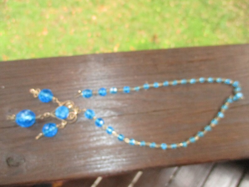May include: A blue beaded necklace with a matching pair of earrings. The necklace is made of small, round beads that are strung together on a thin wire. The earrings are also made of small, round beads that are strung together on a thin wire.