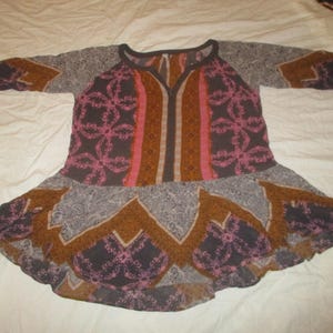 May include: A patterned tunic in shades of brown, pink, and gray, with a flared skirt and long sleeves. The tunic features a V-neck and a floral design. The fabric appears lightweight and suitable for warm weather.