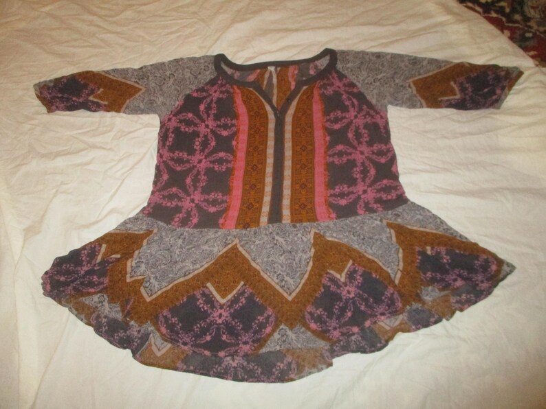 May include: A patterned tunic in shades of brown, pink, and gray, with a flared skirt and long sleeves. The tunic features a V-neck and a floral design. The fabric appears lightweight and suitable for warm weather.