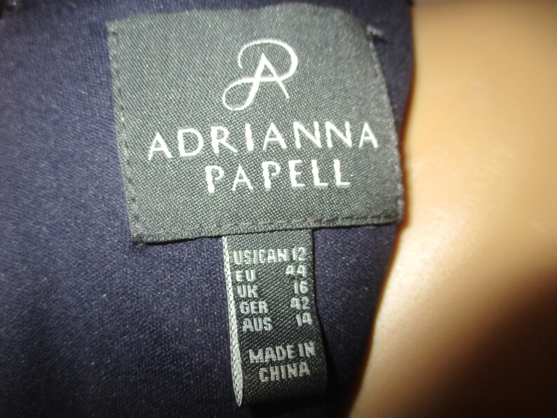 May include: A black label with white text that reads "ADRIANNA PAPELL" and size information: US 12, EU 44, UK 16, GER 42, AUS 14. The label also states "MADE IN CHINA".