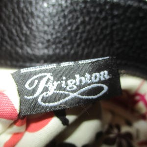 May include: A black fabric label with the word "Brighton" in white cursive script. The label is attached to a black leather bag. The bag's interior lining is visible, with a floral pattern in red and pink.