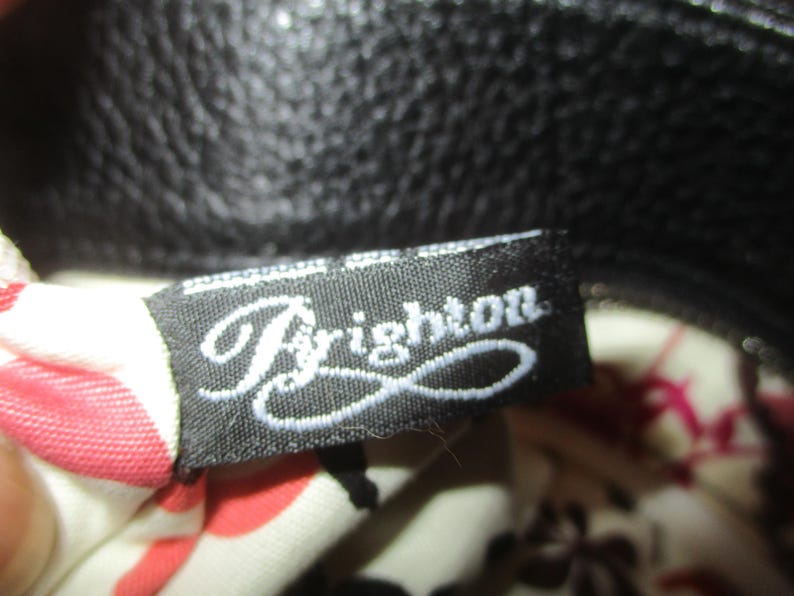 May include: A black fabric label with the word "Brighton" in white cursive script. The label is attached to a black leather bag. The bag's interior lining is visible, with a floral pattern in red and pink.