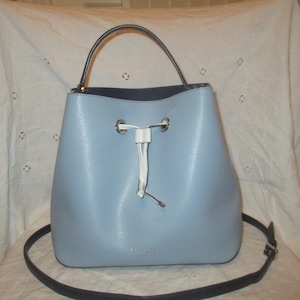 May include: A light blue bucket bag with a textured finish, featuring a single top handle and a long adjustable shoulder strap. The bag has a white drawstring closure threaded through silver-toned metal grommets at the top center.