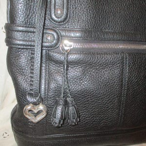 May include: Black leather tote bag with a textured finish. Features a silver zipper, a heart-shaped charm, and two small tassel accents. The bag has a structured design and a strap.