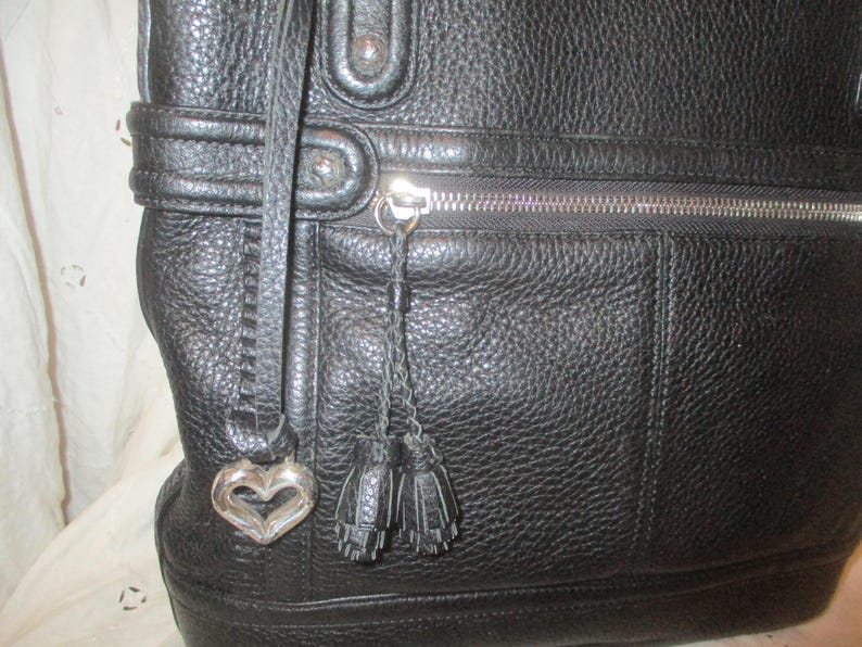May include: Black leather tote bag with a textured finish. Features a silver zipper, a heart-shaped charm, and two small tassel accents. The bag has a structured design and a strap.