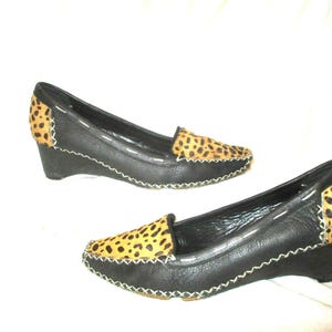 May include: A pair of black leather wedge loafers with leopard print accents on the toe and heel. The shoes have a stitched design around the edges and a small wedge heel.