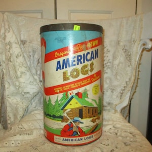 May include: A vintage tin can with a blue, yellow and red label. The label reads "Original American Logs" and features a cartoon illustration of a cabin in the woods. The can is sitting on a white lace tablecloth.
