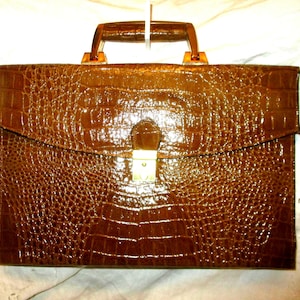 Puede incluir: Un maletín marrón con un patrón texturizado similar al de la piel de cocodrilo. El maletín tiene un cierre de solapa con un cierre dorado y un asa marrón. Una correa de hombro marrón está sujeta a los lados.