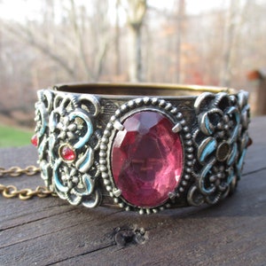 Puede incluir: Un brazalete de plata y oro con una gran pedrería rosa y otras más pequeñas de color rojo y azul. El brazalete tiene una cadena.